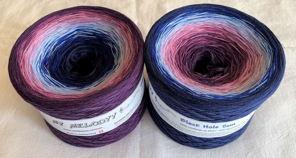 Two cakes of Wolltraum yarn in the colourway Black Hole Sun wound in different directions.  The yarn goes from purple, to light blue, to dark blue.